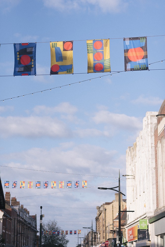 A row of flags hung up along Boscombe high street, the sun is hitting the flags and there are some buildings visible along the highstreet.
