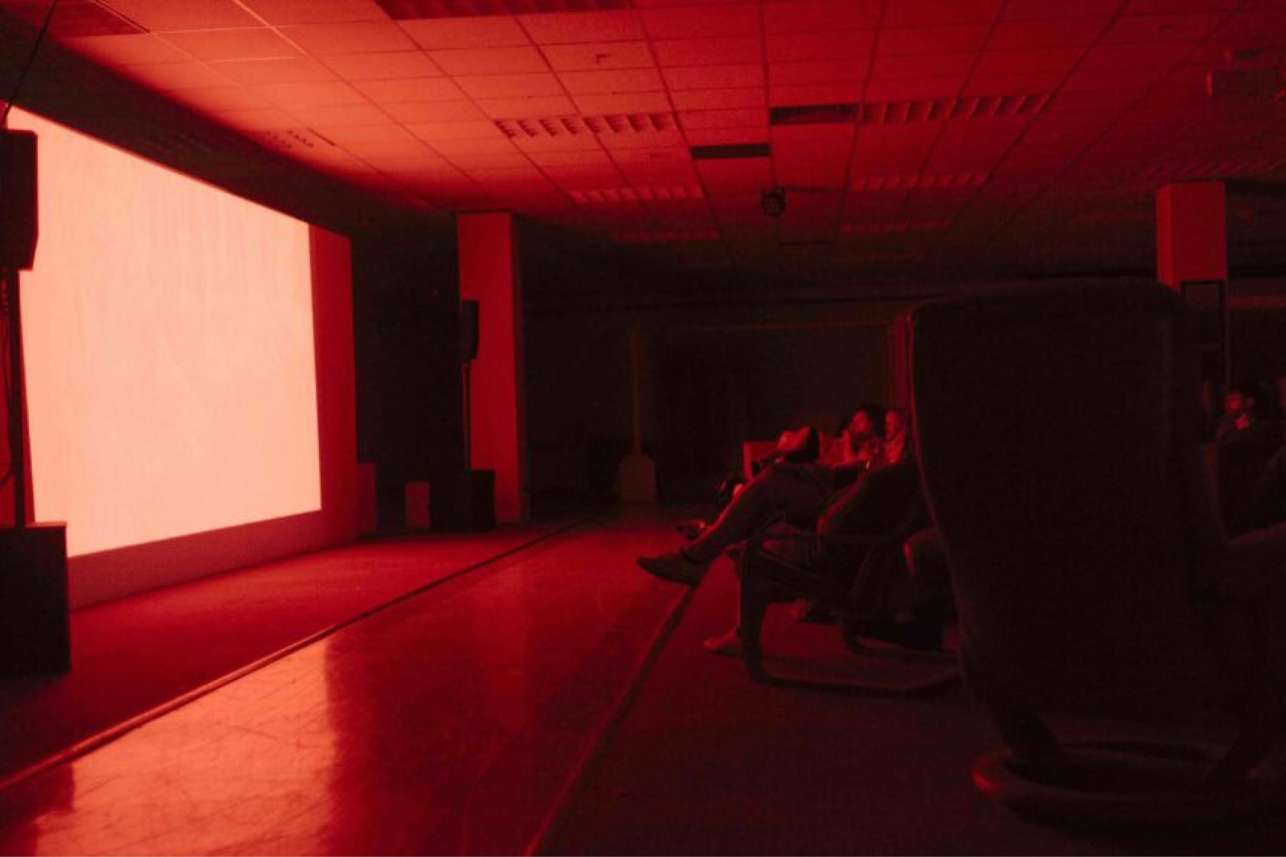 A room filled with red low lit light with a large screen at the front. People are sat in this cinema space looking at the projector and waiting for the film to start.