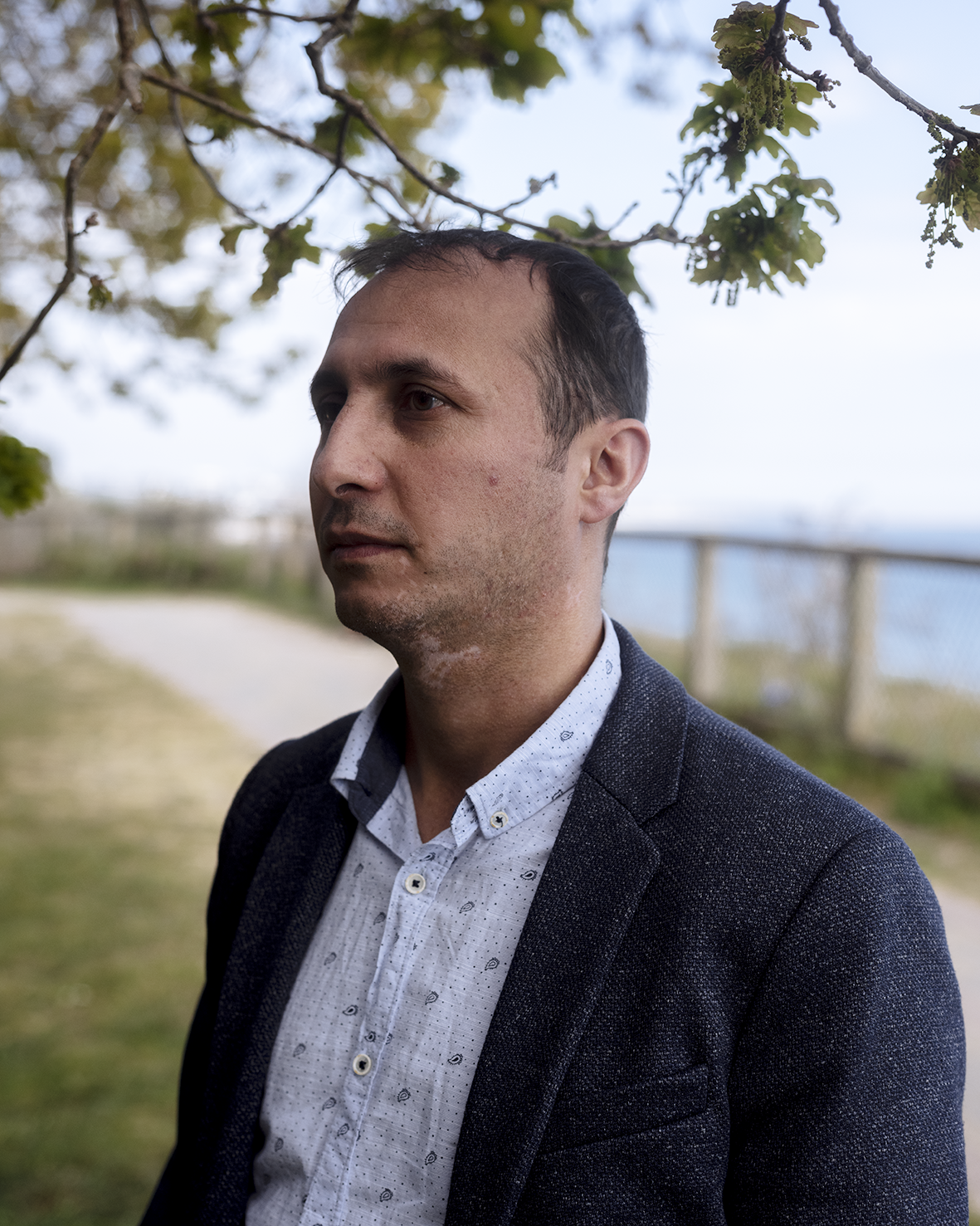 Amir wearing a white short and black jacket, not quite looking at the camera with the sea behind him.