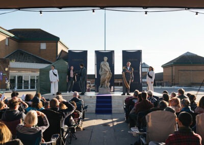 A view from the middle of the performance which has a long purple carpet down the middle leading to the audience. On the stage there is a statue of stone statue and the performers are facing the crowd.