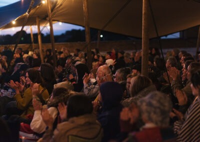 The sun has gone down and the audience are smiling and clapping their hands.