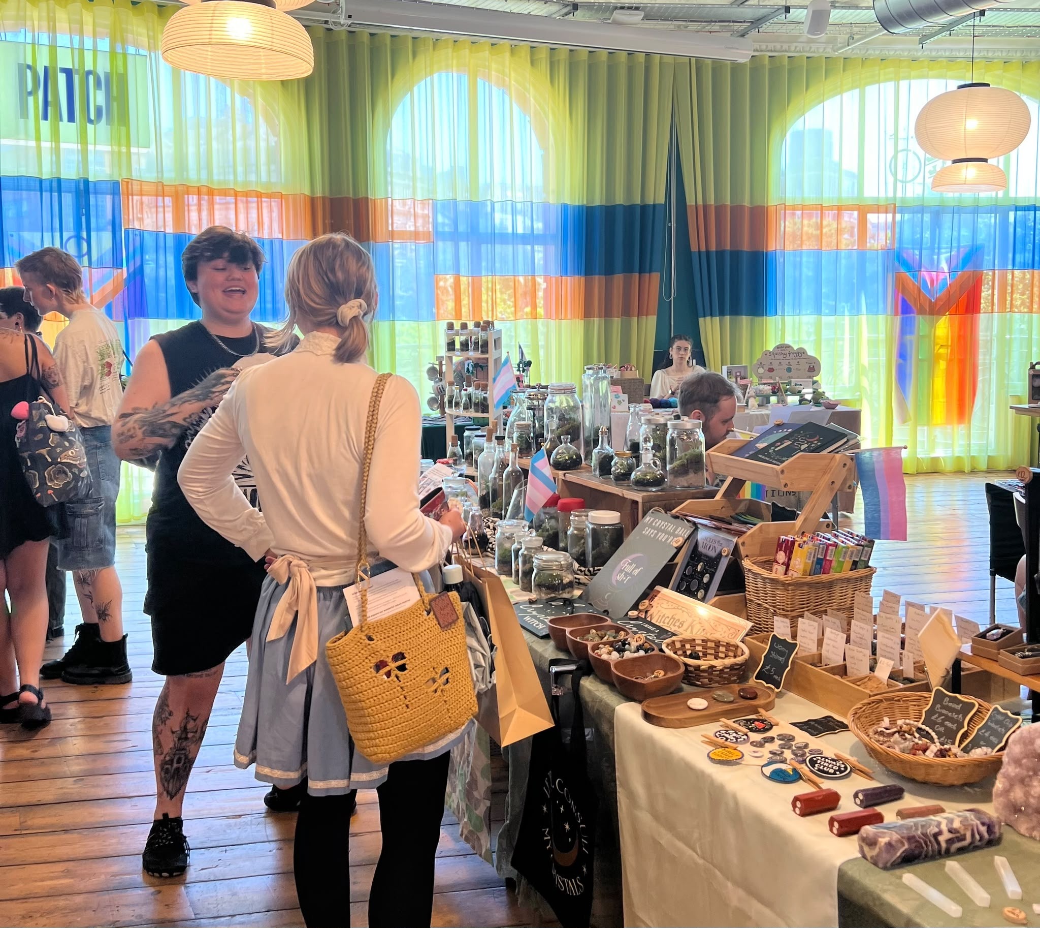 Market stalls set up in a light and bright room, with people talking and laughing while looking at the market stalls